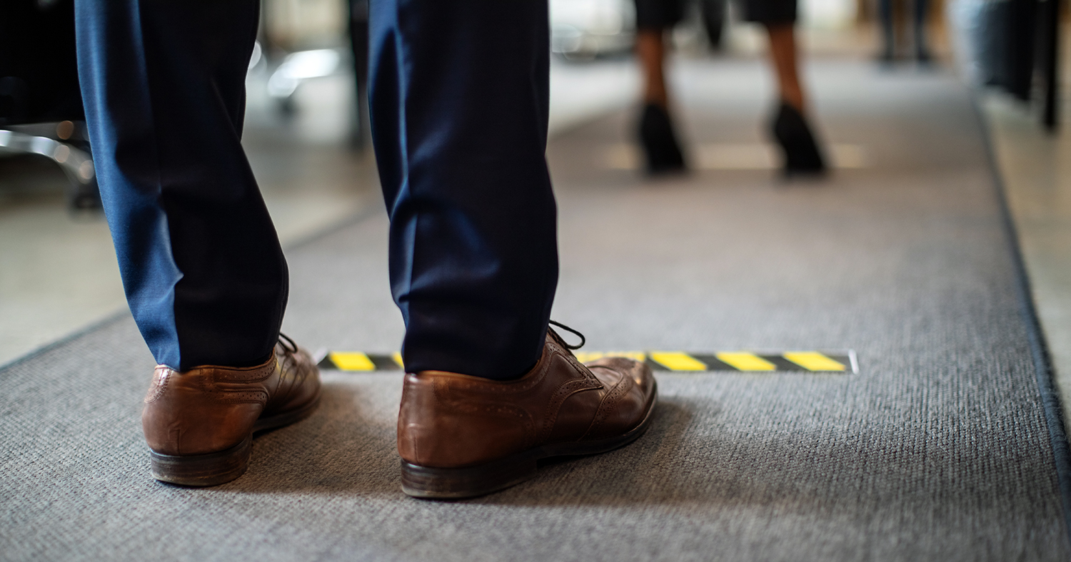 A person stands in a socially distanced line with tape on the floor marking 6 feet between people.