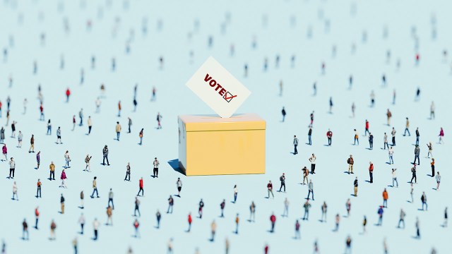 Conceptual image of an oversized voting ballot box in a large crowd of people with shallow depth of field